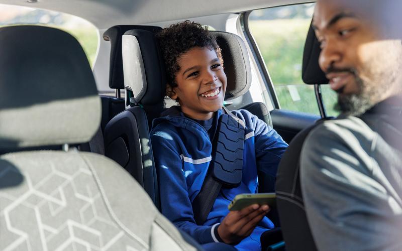 A boy in a car's back seat smiles at a man in the front seat, both wearing seatbelts.