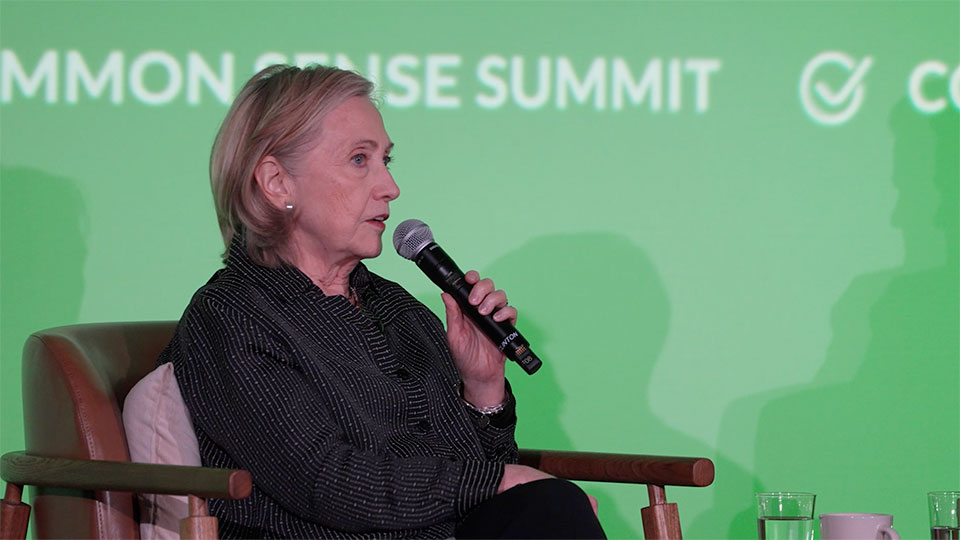 Hillary Clinton holding a mic on stage seated in front of green background